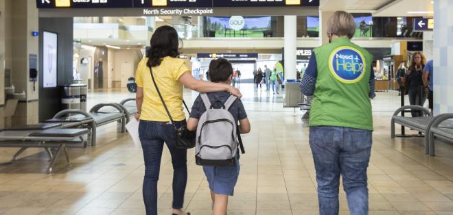 A mother walks with her son through the airport with her arm around him. A helpful volunteer walks with them. 